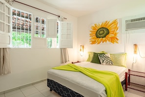 A bedroom with a bed, a wall-mounted light, a window with shutters, and a sunflower painting.