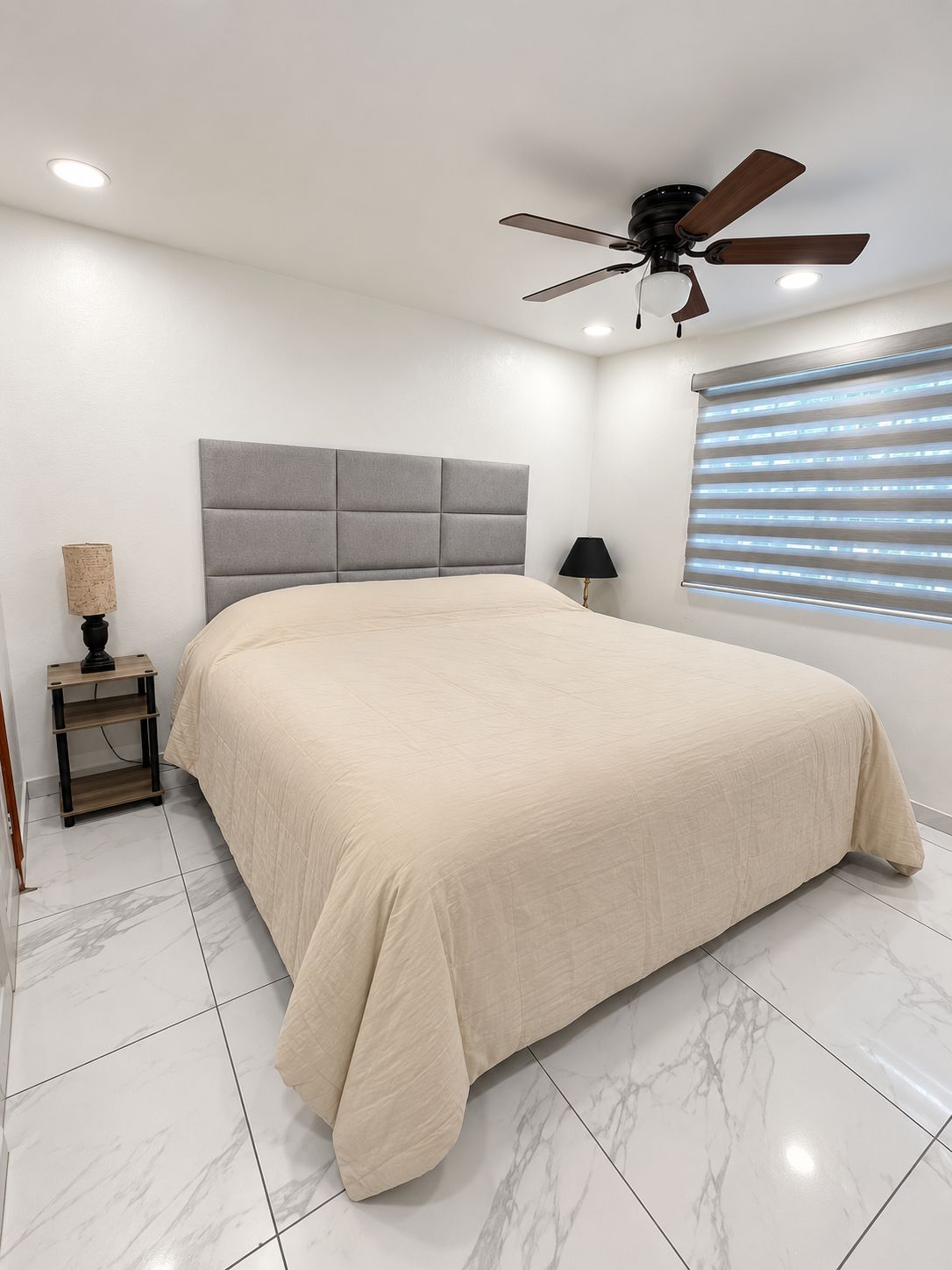 Un dormitorio con cama, ventilador de techo, mesita de noche, lámpara y ventana con persianas.