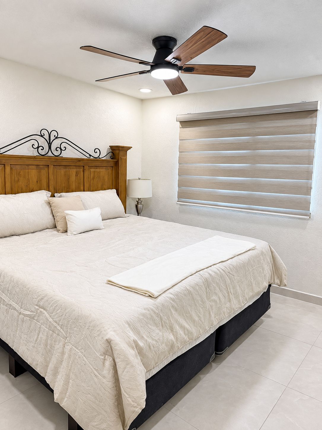 Un dormitorio con una cama, ventilador de techo, lámpara de noche y ventana con persianas.