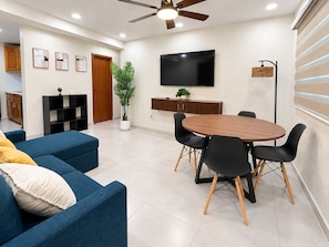 A modern living room with a blue sofa, a wooden dining table with black chairs, a flat-screen TV mounted on the wall, and a ceiling fan.