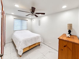 Un dormitorio con cama, ventilador de techo, ventana con persianas, mesita de noche con lámpara y un armario de madera.