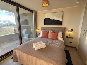 A modern bedroom with a large bed, two bedside lamps, a view through a sliding glass door, and a cityscape visible outside. - Nuevo - Metro - vista a la ciudad  (Santiago)