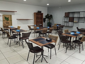 A café with multiple tables and chairs, a wooden bookshelf, and a large potted plant.
