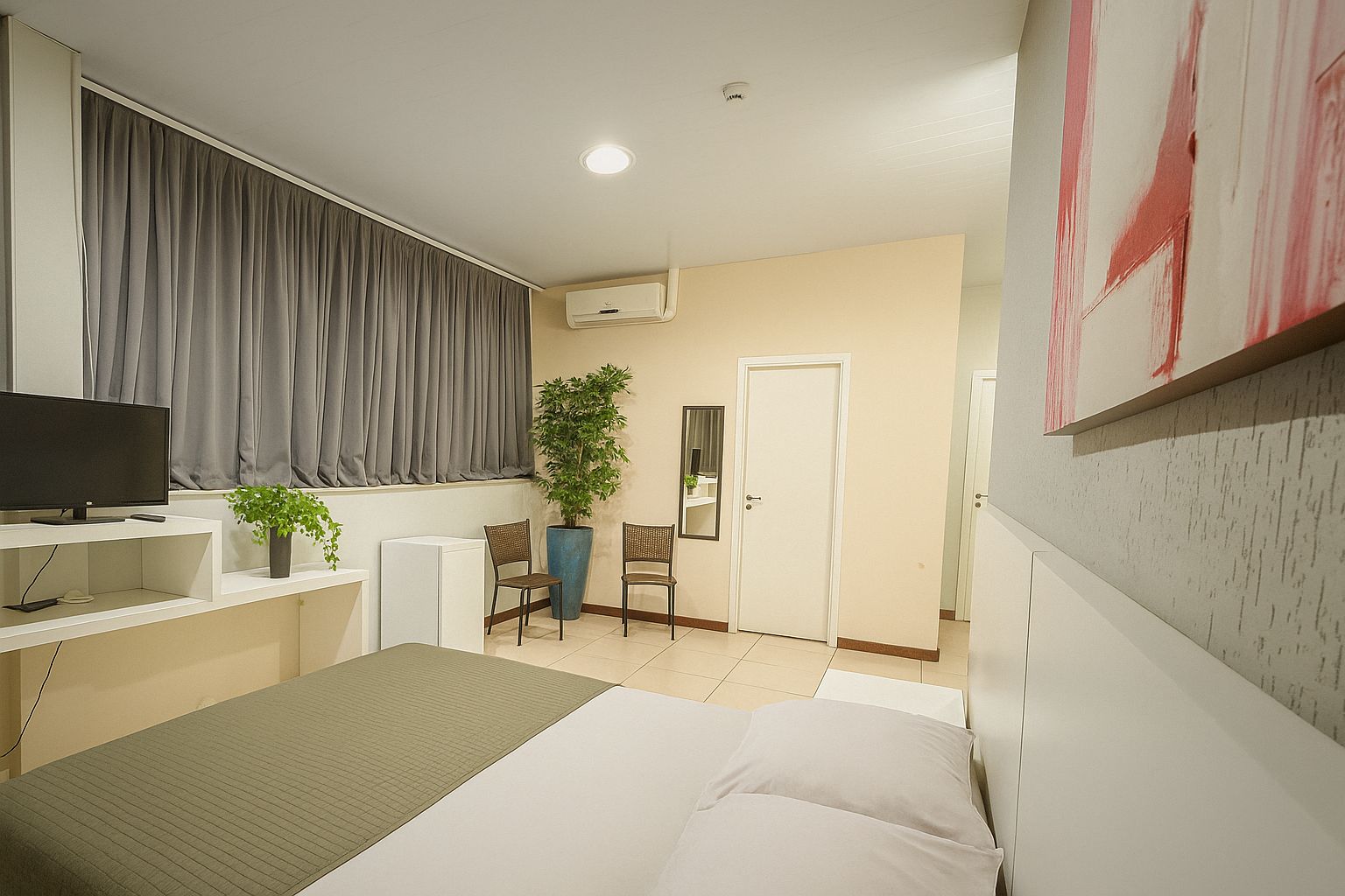 A bedroom with a bed, a desk with a computer, a chair, a potted plant, and a door.
