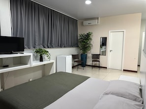 A hotel room with a bed, a desk with a TV, a potted plant, a chair, and a door. - Blu Apart Hotel (Mogi-Guacu)