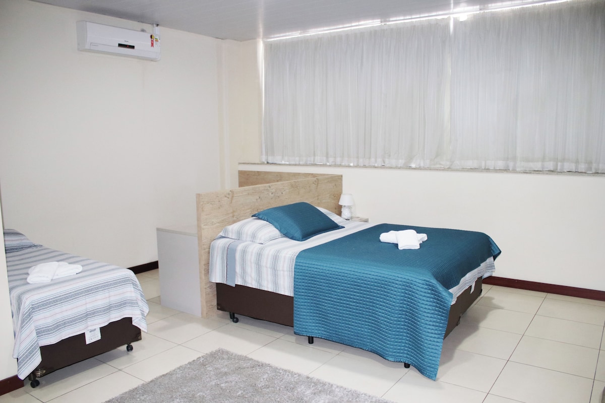 A hotel room with two beds, a small bedside table, a nightstand, a window with curtains, and a wall-mounted air conditioning unit.