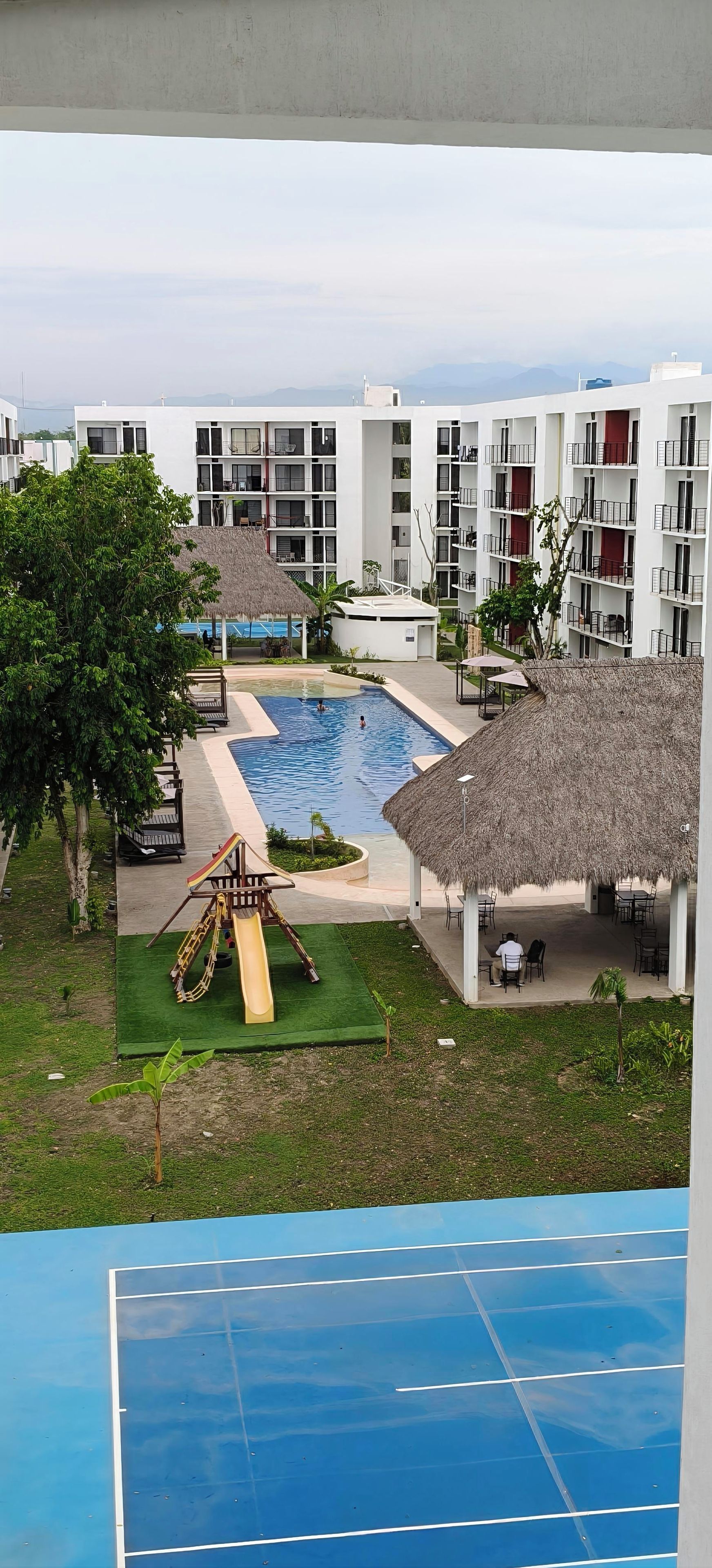 A swimming pool with a slide, surrounded by buildings with balconies, and a tennis court.