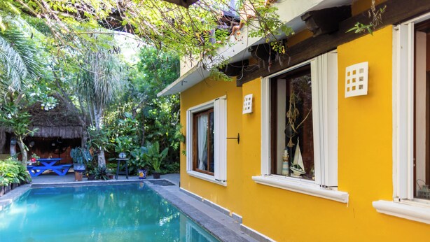 A yellow building with a swimming pool, surrounded by lush greenery and a thatched-roof structure. - A charming villa with access to Yalku Lagoon (Akumal)