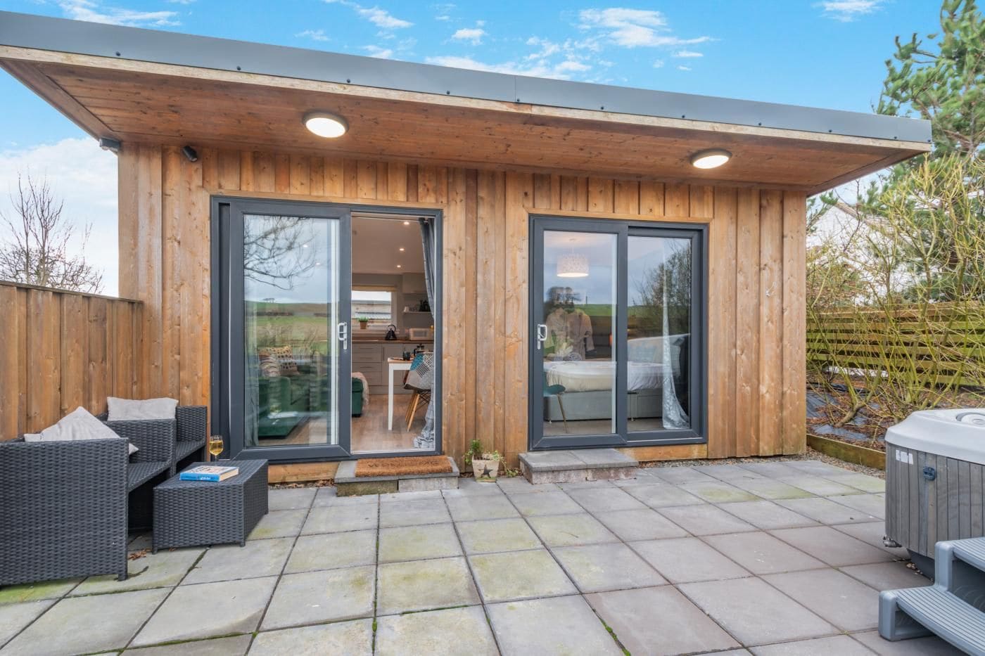 Un bâtiment moderne en bois, doté de grandes portes coulissantes en verre, d’un espace extérieur aménagé et d’un jacuzzi.