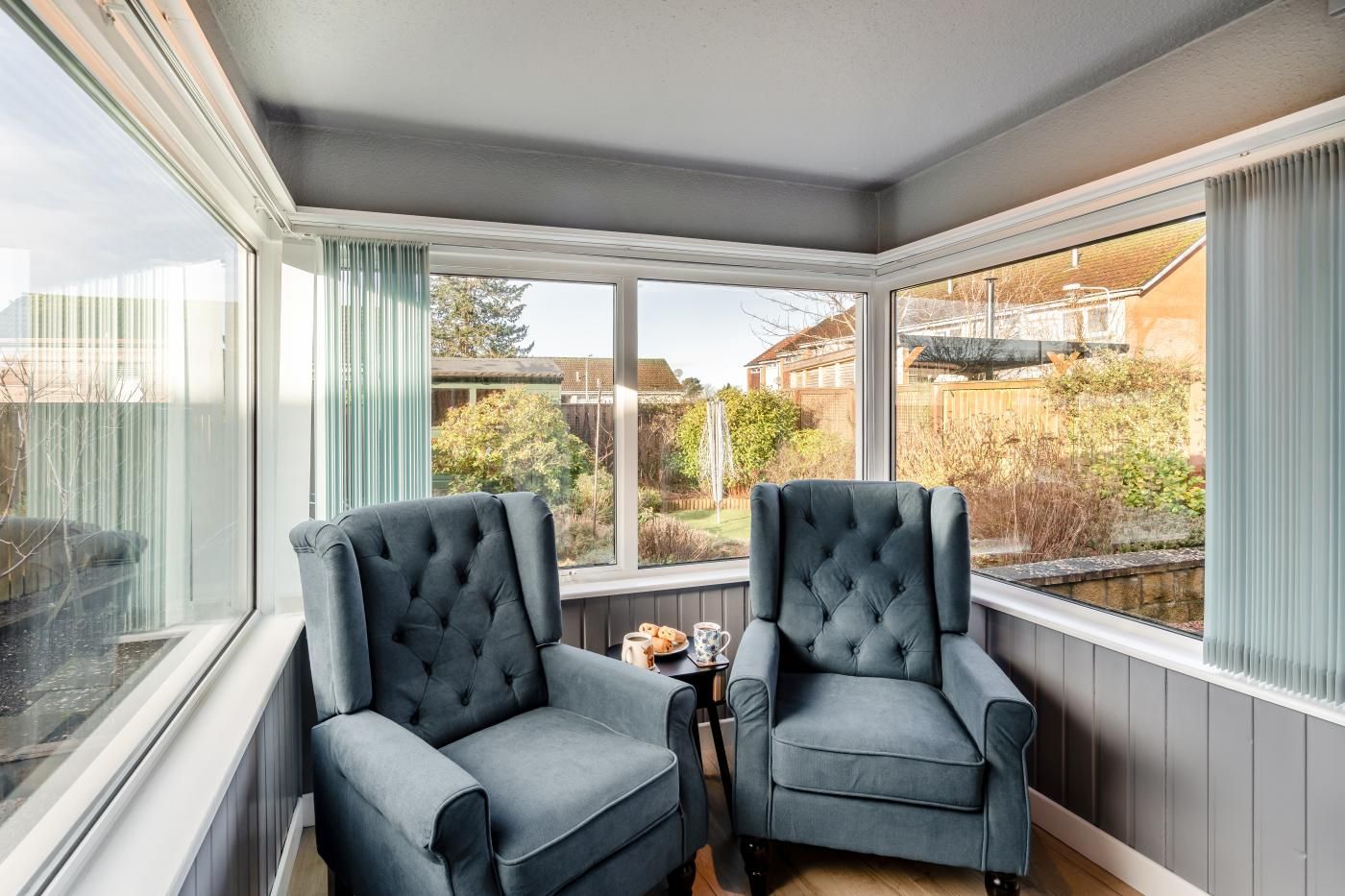 A conservatory with two upholstered armchairs, a small table with a cup and saucer, and a view of a garden and houses.