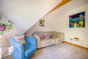 A living room with a blue armchair, a beige sofa, and a glass coffee table. There are two paintings on the walls and a small white table with a bowl of fruit. - 2 Bed in Swanage (oc-t35179) (Swanage)