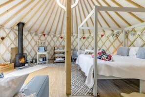 A cozy yurt with a central wood-burning stove, a small table with a lamp, and a comfortable bed with pillows.