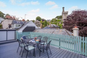 A deck with a table and chairs, surrounded by a light blue fence, overlooking a castle and residential buildings. - 2 Bed in Fowey (oc-s42445) (Fowey)