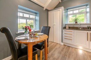 Un coin repas avec une table dressée pour deux personnes, un vase rempli de fleurs et une vue sur l’extérieur grâce à de grandes fenêtres.