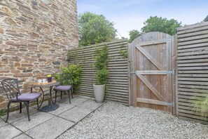 Un mur de pierre, une porte en bois, une table avec des chaises et des plantes en pot dans une cour.