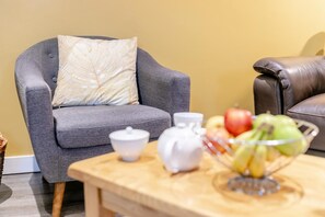 A living room with a grey armchair, a brown leather sofa, a wooden coffee table with a fruit bowl, a white teapot, and a white cup. - 1 Bed in Duloe (oc-h27527) (Duloe)