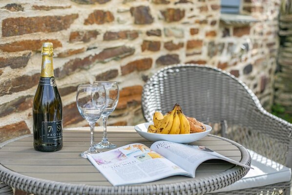Una botella de Prosecco, dos copas de vino, un plato con plátanos y un muffin, y una revista sobre una mesa de mimbre al aire libre.