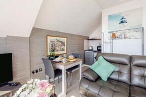 Ein modernes Wohnzimmer mit einem braunen Ledersofa, einem Esstisch mit Frühstücksgebäck, einem Flachbildfernseher und einem Gemälde eines Leuchtturms.