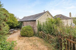 Een stenen huis met een schuin dak, een omheinde tuin en een grindpad dat leidt naar een terras met tuinmeubilair.