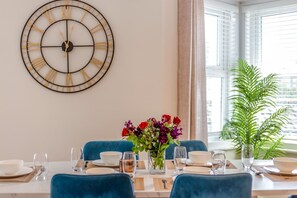 Ein Essbereich mit einer runden Wanduhr, einem für zwei Personen gedeckten Tisch mit blauen Stühlen und einem Fenster mit Blick auf Grünflächen.