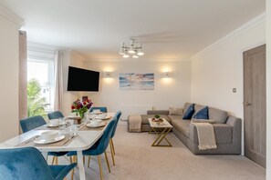 A modern living room with a dining area, a grey sofa, blue chairs, and a flat-screen TV mounted on the wall. - 3 Bed in Bournemouth (oc-s44899) (Bournemouth)