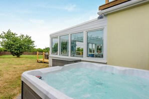 A hot tub adjacent to a building with large windows, a wooden deck, and a tree in the background. - 6 Bed in Moreton (oc-h45423) (Moreton)