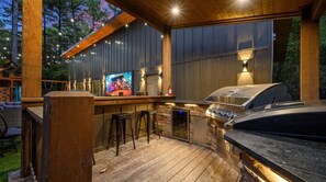 An outdoor bar area with a grill, bar stools, and a television screen. - The Cedarwood Collective · Indoor Pool, Theatre, Putt-Putt & Gameroom | Sleeps 27! (Broken Bow)