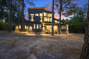 A modern two-story building with a gravel driveway and a wooden deck, surrounded by trees. - The Cedarwood Collective · Indoor Pool, Theatre, Putt-Putt & Gameroom | Sleeps 27! (Broken Bow)
