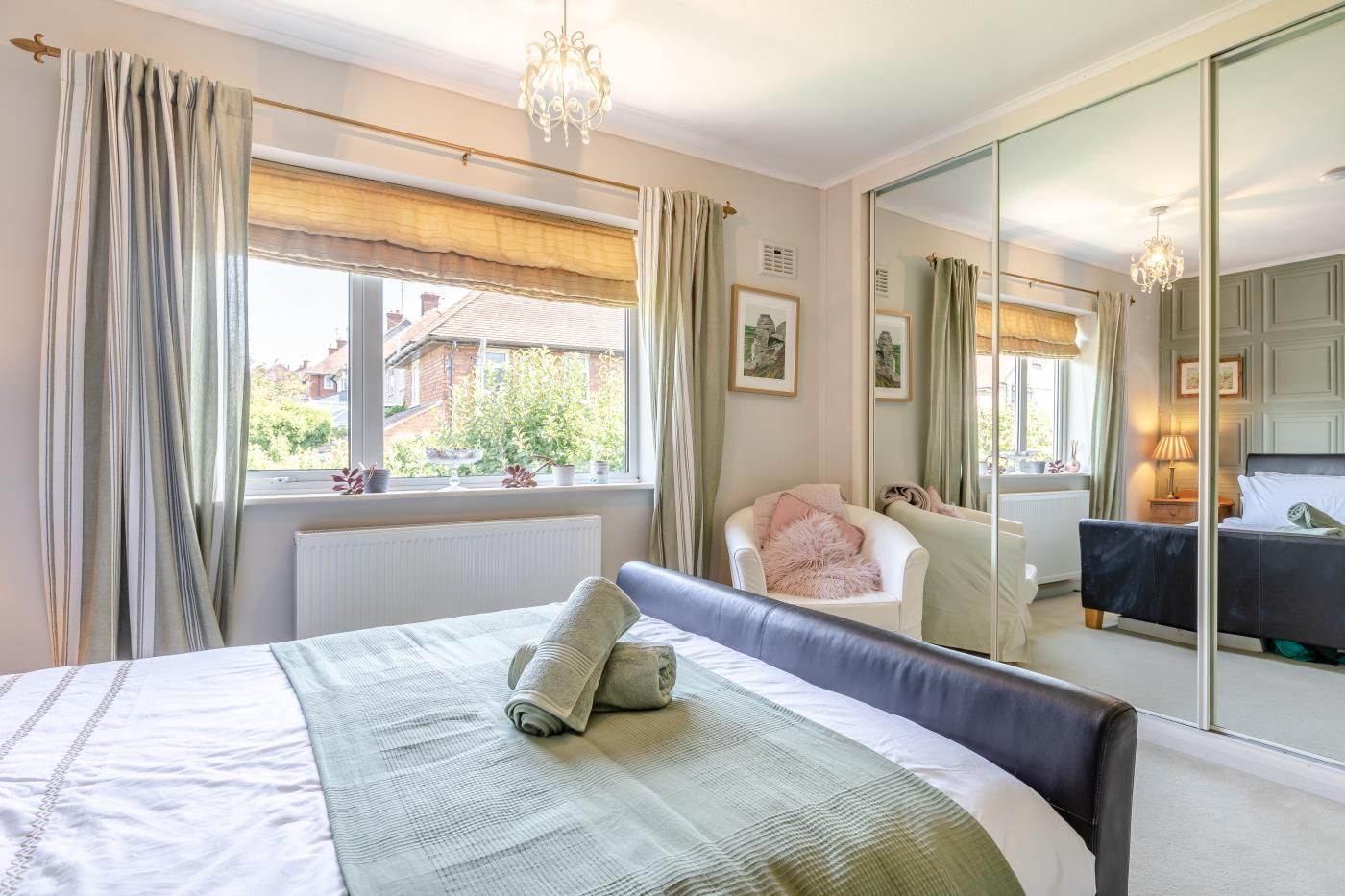 A bedroom with a bed, a chair, a large mirror, and a window with curtains.