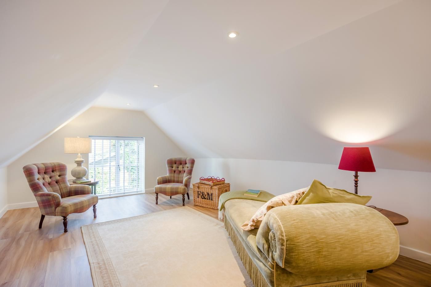 A spacious attic bedroom with a high ceiling, a large window, a sofa, two armchairs, a lamp, and a basket.