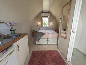 Ein kleines Zimmer mit Küchenzeile, einem Bett mit Kopfteil, einem Spiegel und einem fenster mit Vorhängen.