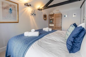 Ein ordentlich gemachtes Bett mit blauer Decke und weißen Kissen, ein Holzregal mit verschiedenen Gegenständen und ein gerahmtes Kunstwerk an der Wand.