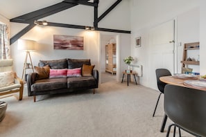 Ein Wohnzimmer mit einer Couch, einem Esstisch und einem Sessel. Eine Lampe, eine Pflanze und ein Gemälde sind an der Wand zu sehen.