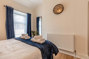 A bedroom with a bed, two towels, a clock, a window with curtains, a radiator, and a mirror. - 2 Bed in Buxton (oc-p45950) (Buxton)