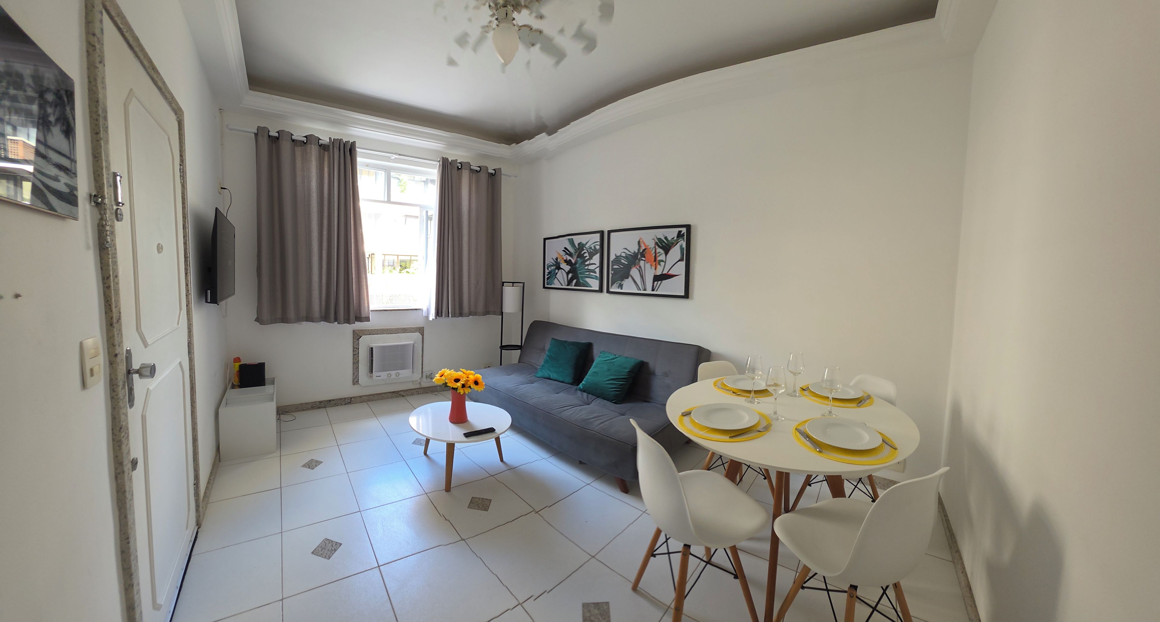 Ein Wohnzimmer mit einer Couch, einem Esstisch gedeckt mit Tellern und Gläsern, einer Vase mit Blumen und einem Fernseher an der Wand.