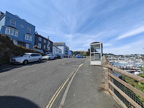 Une route côtière bordée de voitures garées, un arrêt de bus et une vue sur un port de plaisance.