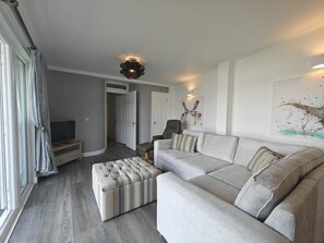 A modern living room with a sectional sofa, a tufted ottoman, a television, and artwork on the walls. - 1 Bed in Kingswear (oc-b45934) (Dartmouth)