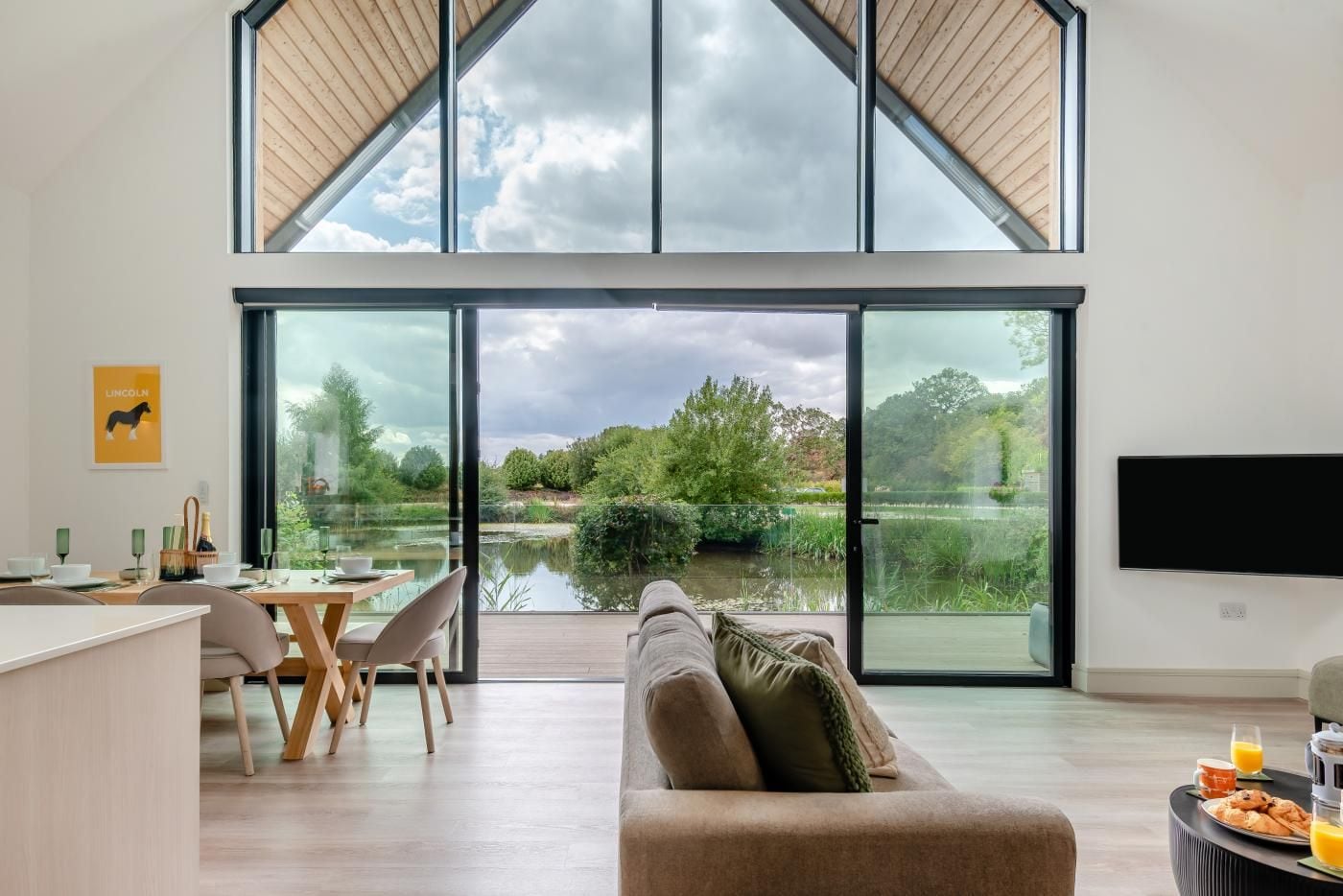 A modern living room with a large glass door leading to a garden view, a dining area with a table and chairs, and a flat-screen TV mounted on the wall.