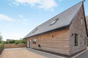 A modern wooden house with a sloped roof, a small porch, and a gravel driveway. - 3 Bed in Doddington (oc-l45736) (Doddington)