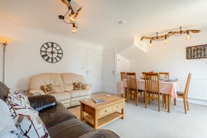 A living room with a sofa, a coffee table, a dining area with a table and chairs, and a wall clock. - 3 Bed in Westward Ho (oc-h45910) (Northam)
