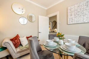 A dining area with a glass table set for four, a sofa with cushions, and a door leading to a kitchen. - 2 Bed in Scarborough (oc-c45973) (Scarborough)