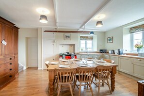 A dining area with a wooden table set for six, a kitchen in the background, and a window with a view. - 4 Bed in Ebberston (oc-h45939) (Ebberston)
