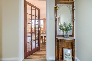 Um corredor com porta de madeira que dá para uma sala de jantar, um espelho com moldura decorativa e um vaso com flores sobre um aparador.
