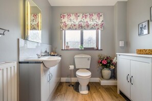 Ein Badezimmer mit weißem Waschbecken, Toilette und Schrank, einem Fenster mit floralem Vorhang und einem Spiegel.