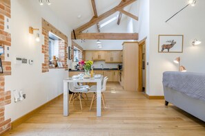 Eine moderne Küche mit Holzbalken, ein Essbereich mit weißem Tisch und Stühlen sowie ein Bett mit grauer Decke.