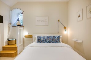 Ein Schlafzimmer mit einem Bett, einem Nachttisch, einer Lampe, einem an der Wand befestigten Regal, einem gerahmten Bild und einer Treppe mit einem Korb.