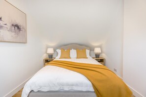 Ein ordentlich bezogenes Bett mit weißer Daunendecke, zwei Kissen, einer senfgelben Tagesdecke und zwei Nachttischlampen auf Holznachttischen.