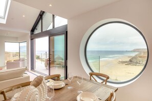 A modern interior with a dining area, a large round window with a beach view, and a glass door leading to an outdoor deck. - 4 Bed in Porthtowan (oc-m45962) (Porthtowan)