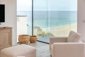 Ein modernes Wohnzimmer mit großem Fenster mit Blick auf einen Strand. Auf dem Boden stehen zwei Körbe aus Weidengeflecht, ein an der Wand befestigtes Fernsehgerät und ein gestreiftes Sofa.