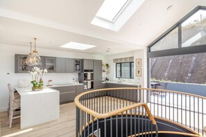 A modern kitchen with a central island, a dining area with a white table, and a staircase leading to an outdoor balcony. - 4 Bed in Porthtowan (oc-45963) (Porthtowan)